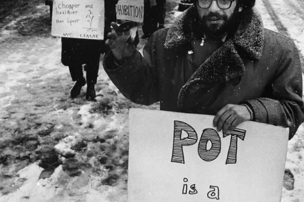 Black-and-white photograph of poet Allen Ginsberg standing in the snow holding a handmade POT IS A REALITY KICK sign, surrounded by marijuana legalization protesters.