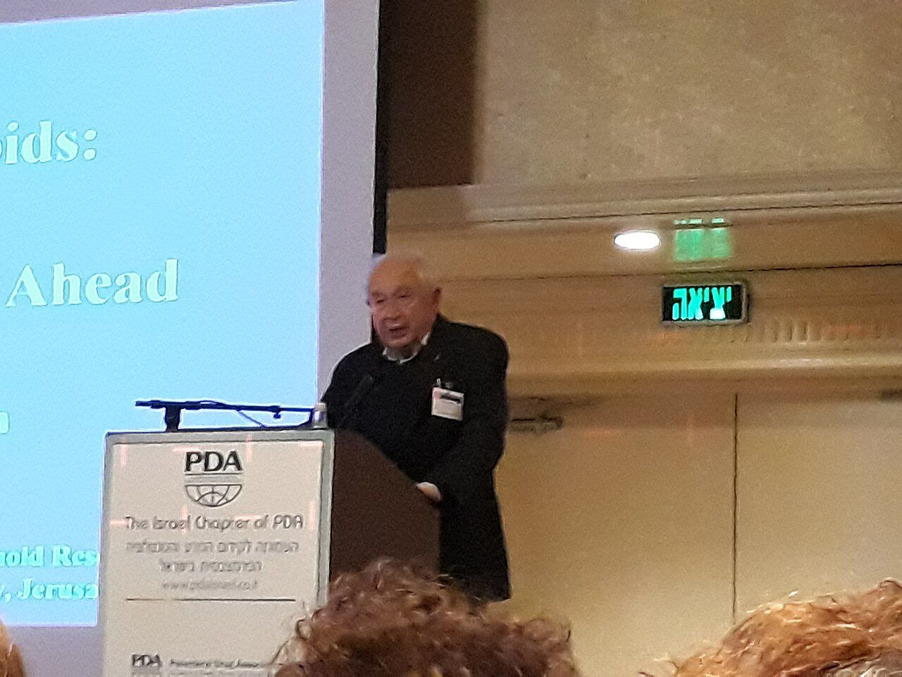 An elderly man speaking at a lectern in front of a projected slide