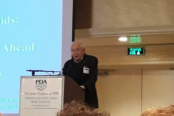 An elderly man speaking at a lectern in front of a projected slide