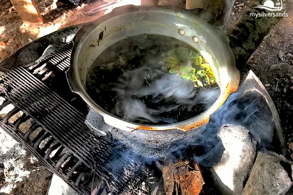 Documentary photograph of a steaming metal pot filled with green leaves over a rustic wood fire at a Jamaican Rastafari farmer's home.