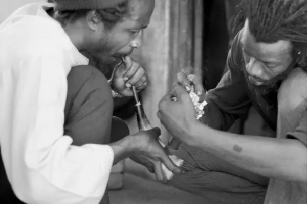 Historical photograph of two Rastafari participants lighting and smoking a communal cannabis chalice during a religious ceremony in Kingston.