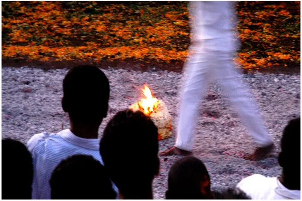 Silhouetted spectators watch a man in white walk across a bed of embers, with flowers banked behind
