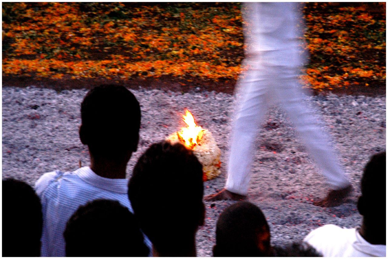 Silhouetted spectators watch a man in white walk across a bed of embers, with flowers banked behind