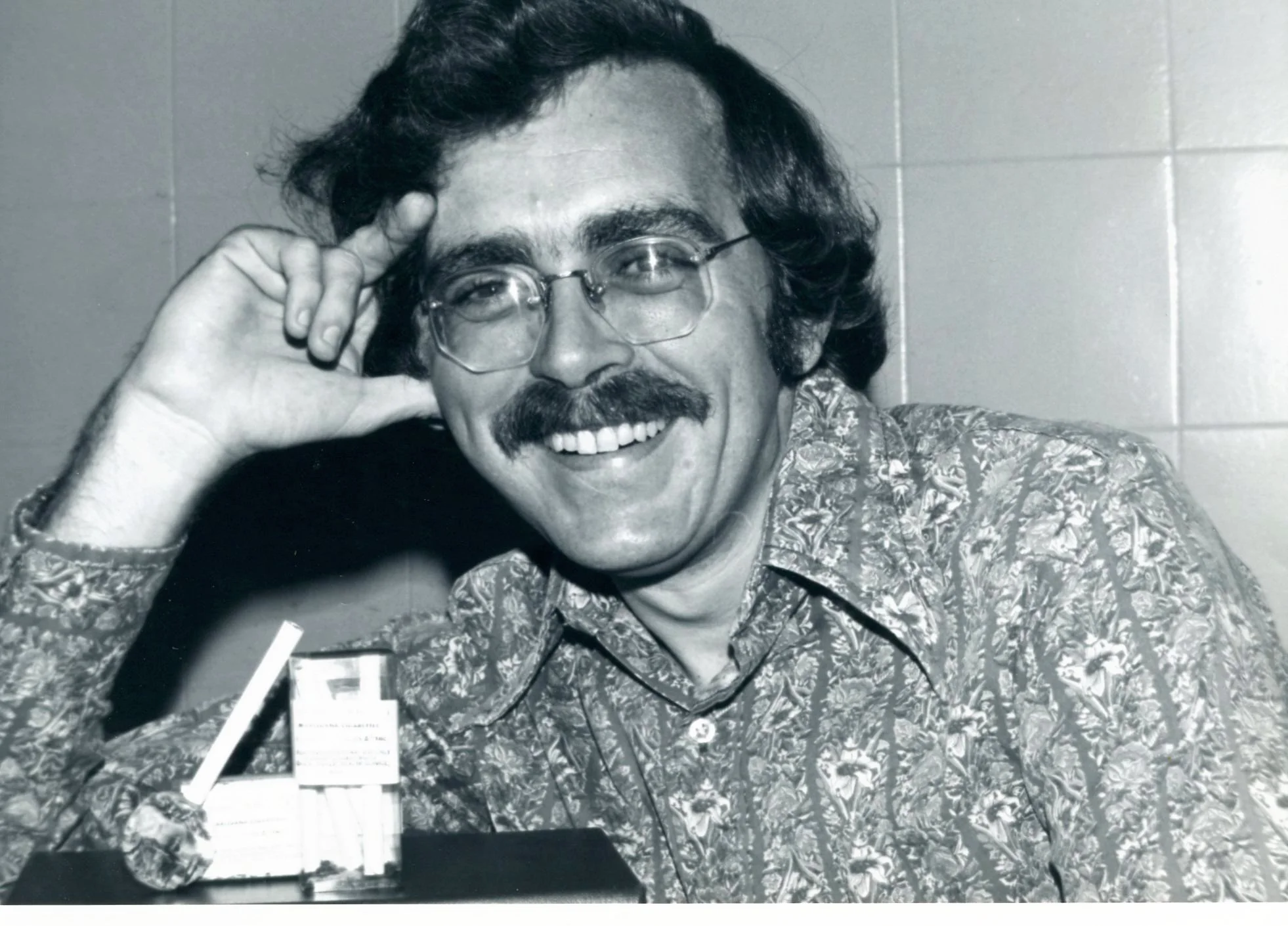Black-and-white photograph of Robert Randall smiling in glasses beside his first legal supply of government-provided marijuana cigarettes in November 1976.