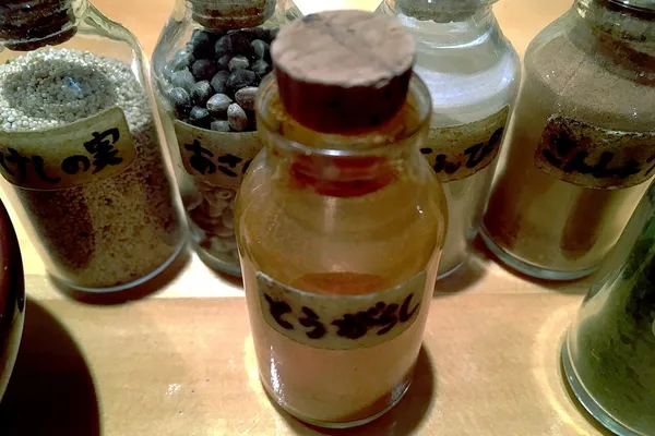 Small glass jars labelled in Japanese, holding poppy seed, whole hemp seeds, ground chili, dried citrus peel and sansho pepper
