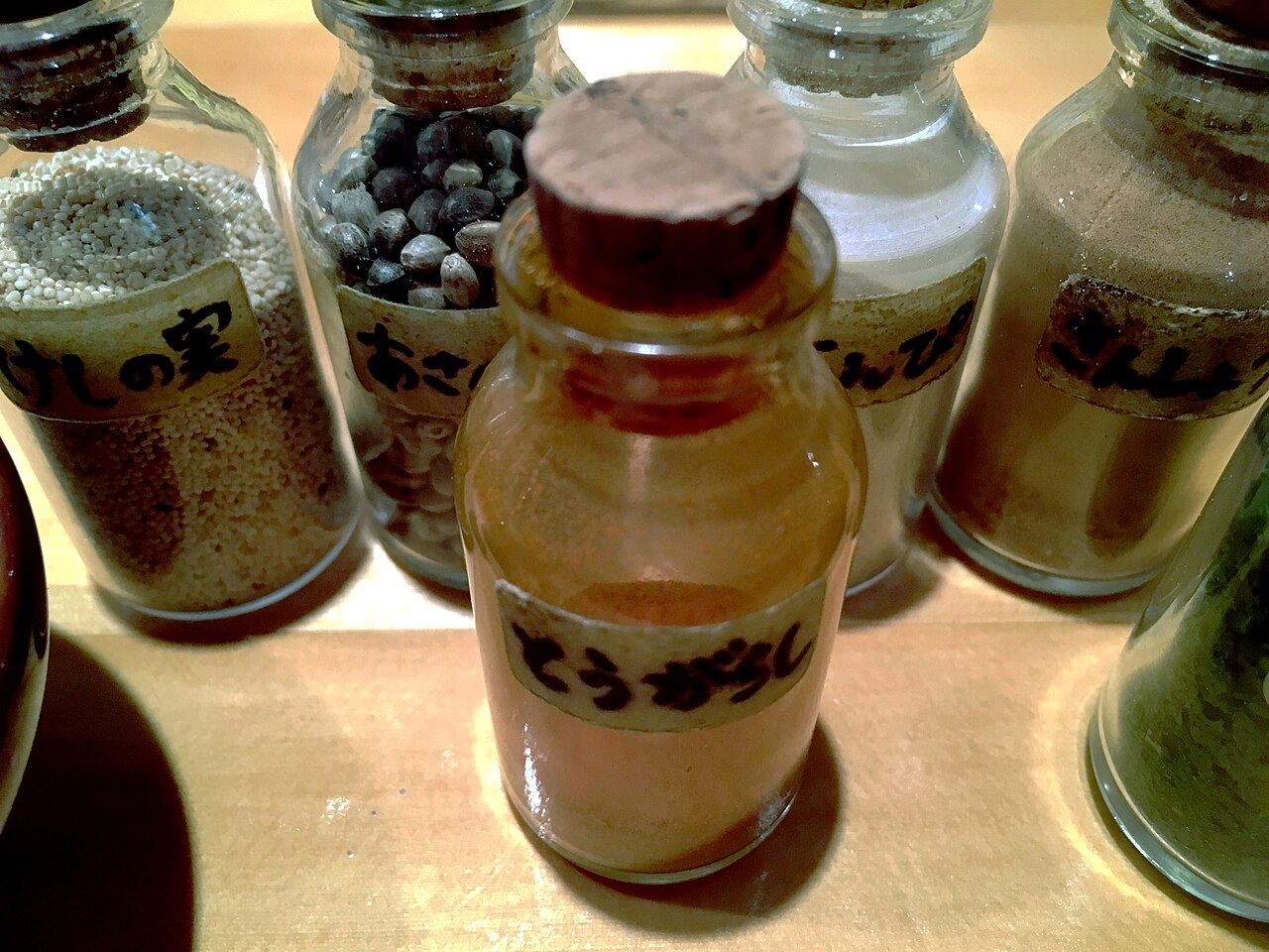 Small glass jars labelled in Japanese, holding poppy seed, whole hemp seeds, ground chili, dried citrus peel and sansho pepper