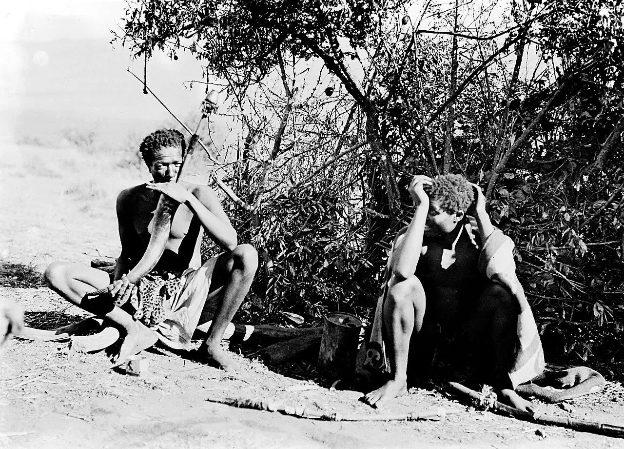 A young man seated beside a bush smoking a long horn water pipe; a second man sits nearby with his head in his hands