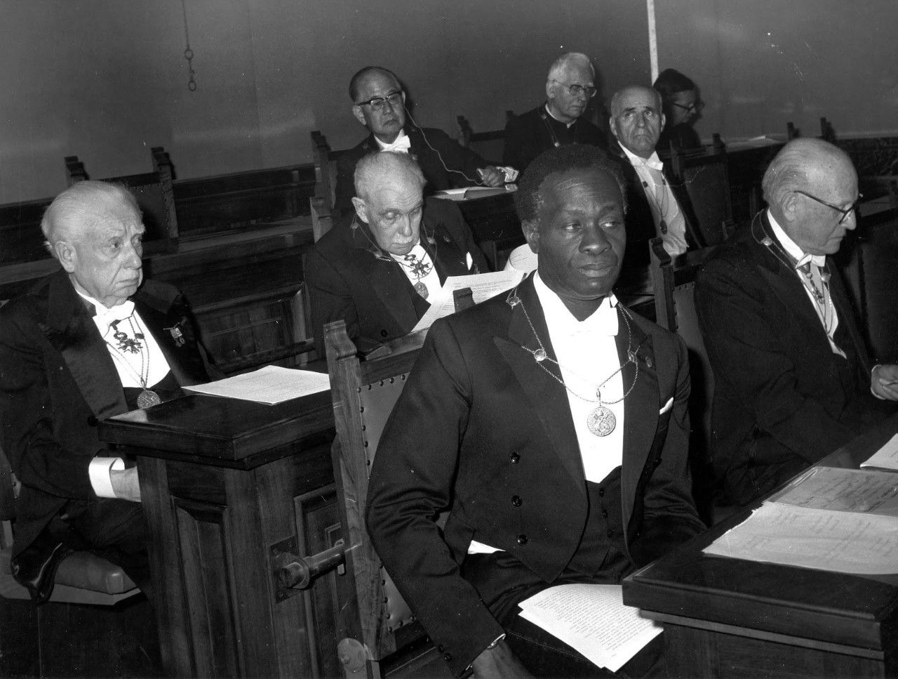Historical black-and-white photograph of Nigerian psychiatrist Thomas Adeoye Lambo seated at an academic gathering in 1975, wearing a ceremonial medallion.