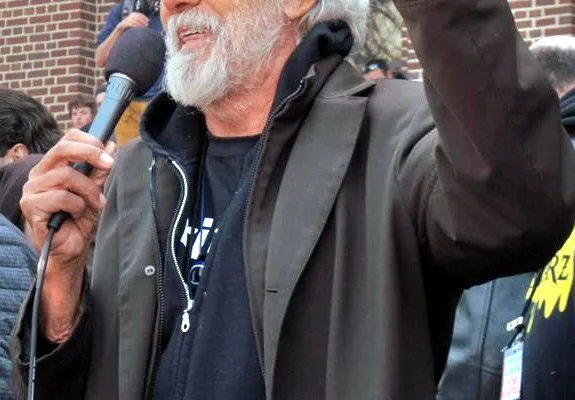 Tommy Chong holding a microphone and raising his hand while speaking to a crowd outdoors.