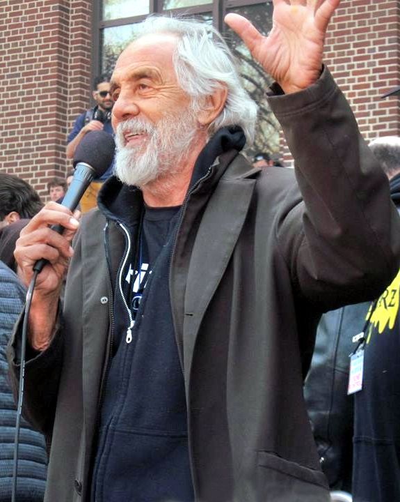 Tommy Chong holding a microphone and raising his hand while speaking to a crowd outdoors.