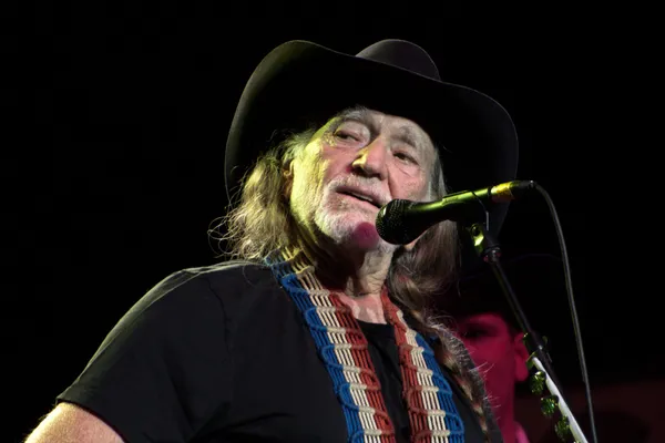 Willie Nelson in a black cowboy hat singing into a microphone on stage.