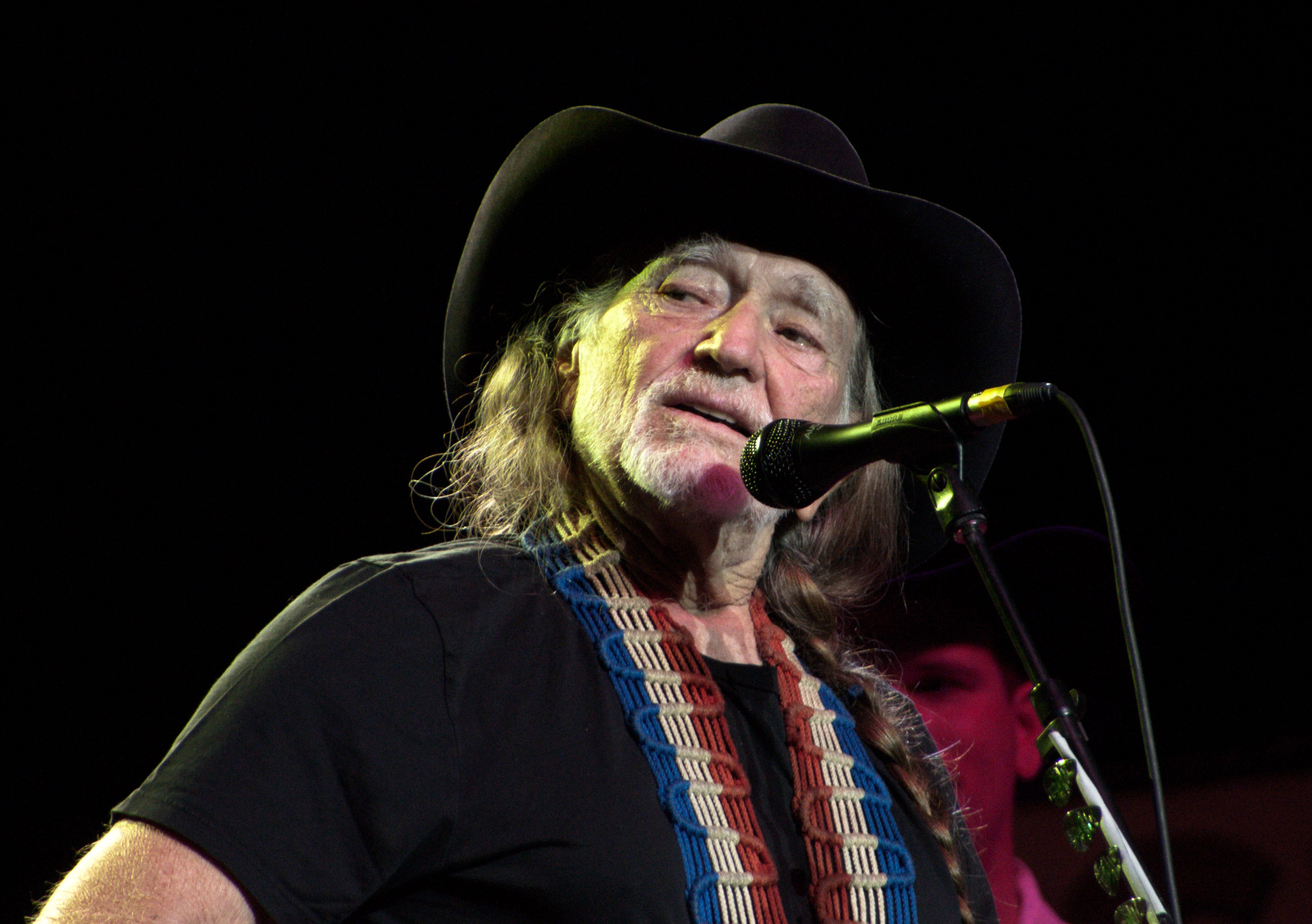 Willie Nelson in a black cowboy hat singing into a microphone on stage.